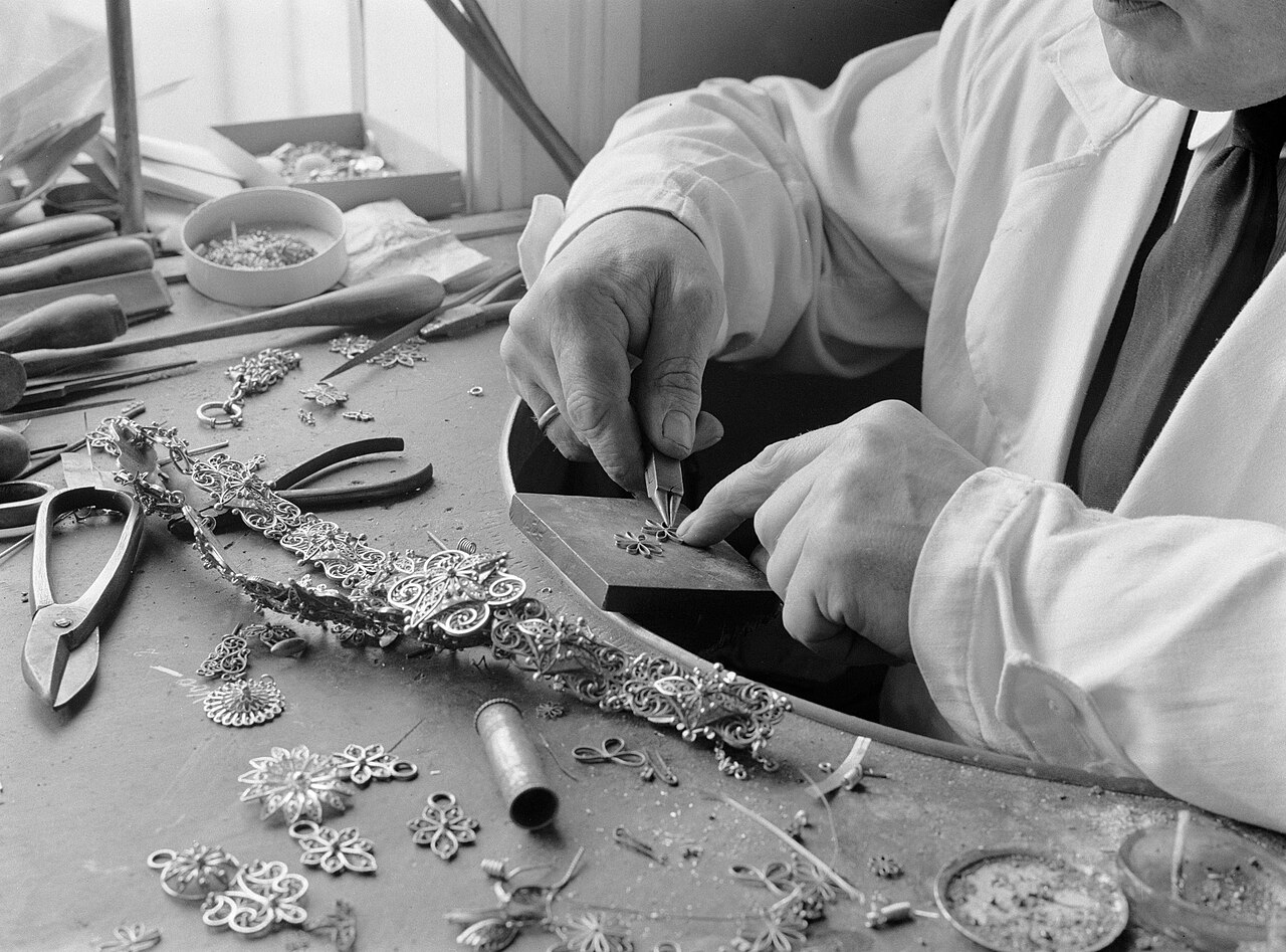 A black-and-white photo of a goldsmith working at a bench covered with tools and jewelry parts